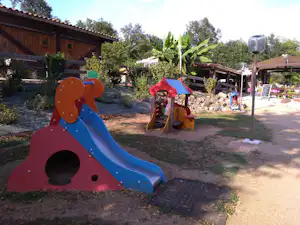 Bunter Spielplatz mit Rutsche und Kletterelementen auf dem Campingplatz, im Hintergrund Holzhäuser und Bäume.