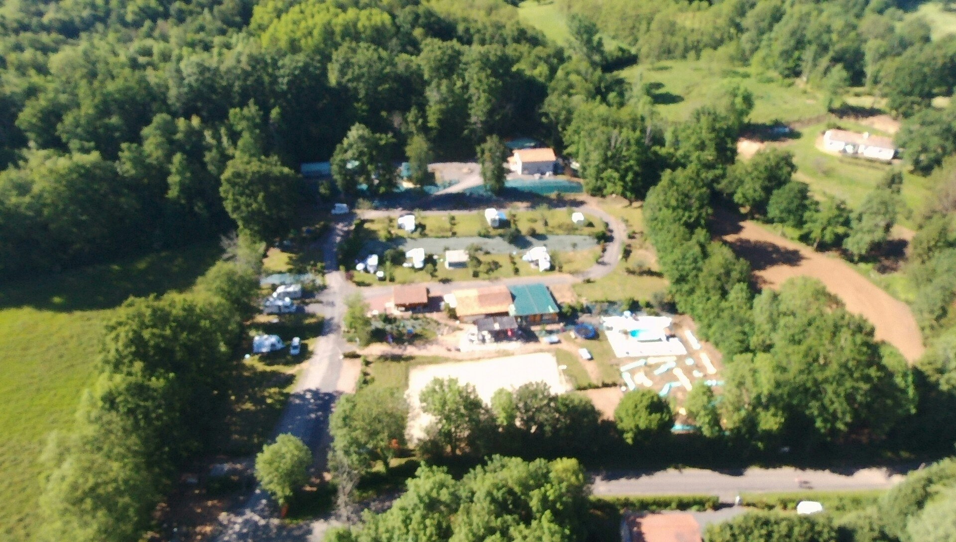 Luftaufnahme eines Campingplatzes mit Gebäuden, Stellplätzen und Wegen im Grünen, umgeben von Bäumen und Wiesen.