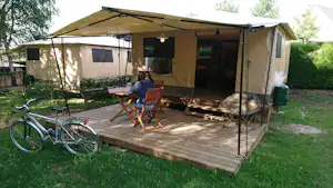 Campingzelt mit überdachter Terrasse, Holzpodest und Stühlen; daneben ein Fahrrad im grünen Garten.