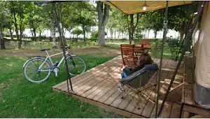 Holzdeck am Campingplatz mit Campingstuhl und Fahrrad im Grünen, umgeben von Bäumen und Zeltüberdachung.