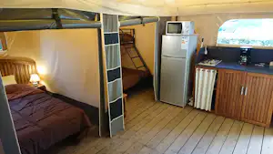 Innenansicht eines Camping-Mobilheims mit Bett, Holzfußboden, Küchenzeile mit Kühlschrank und Schrank sowie Vorhang am Eingang.