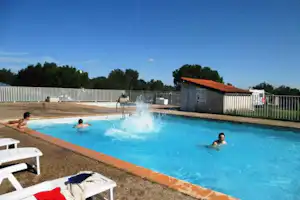 Camping-Pool mit blauem Wasser, Sonnenliegen am Beckenrand und Zaun, im Hintergrund ein Gebäude mit rotem Dach.