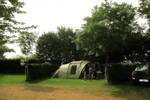 Zelt auf einem Campingplatz im Grünen, mit Sitzgruppe und Auto am Rand, umgeben von Hecken und Bäumen.