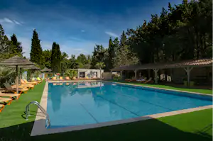 Schwimmbecken eines Campingplatzes mit Liegen, Sonnenschirmen und überdachten Bereichen, umgeben von Bäumen und Rasen.