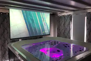 Whirlpool im Wellnessraum mit beleuchtetem Wasser, gepolsterten Wänden und einer Leinwand im Hintergrund.