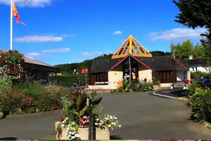 Campingplatz-Gebäude mit Giebeldach und Holzverkleidung, Blumenbeete und Einfahrt, im Hintergrund Bäume und Himmel.