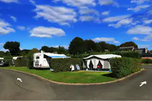 Wohnwagen und Zelt auf einem Campingplatz, mit Hecken, Rasenfläche und Parkplatz am Straßenrand.