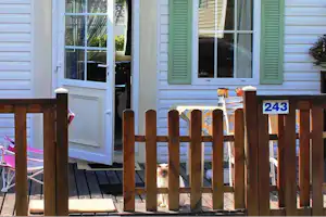 Campinghaus mit grünen Fensterläden, Holzterrasse und Zaun; im Hintergrund ist eine Katze vor der Tür zu sehen.