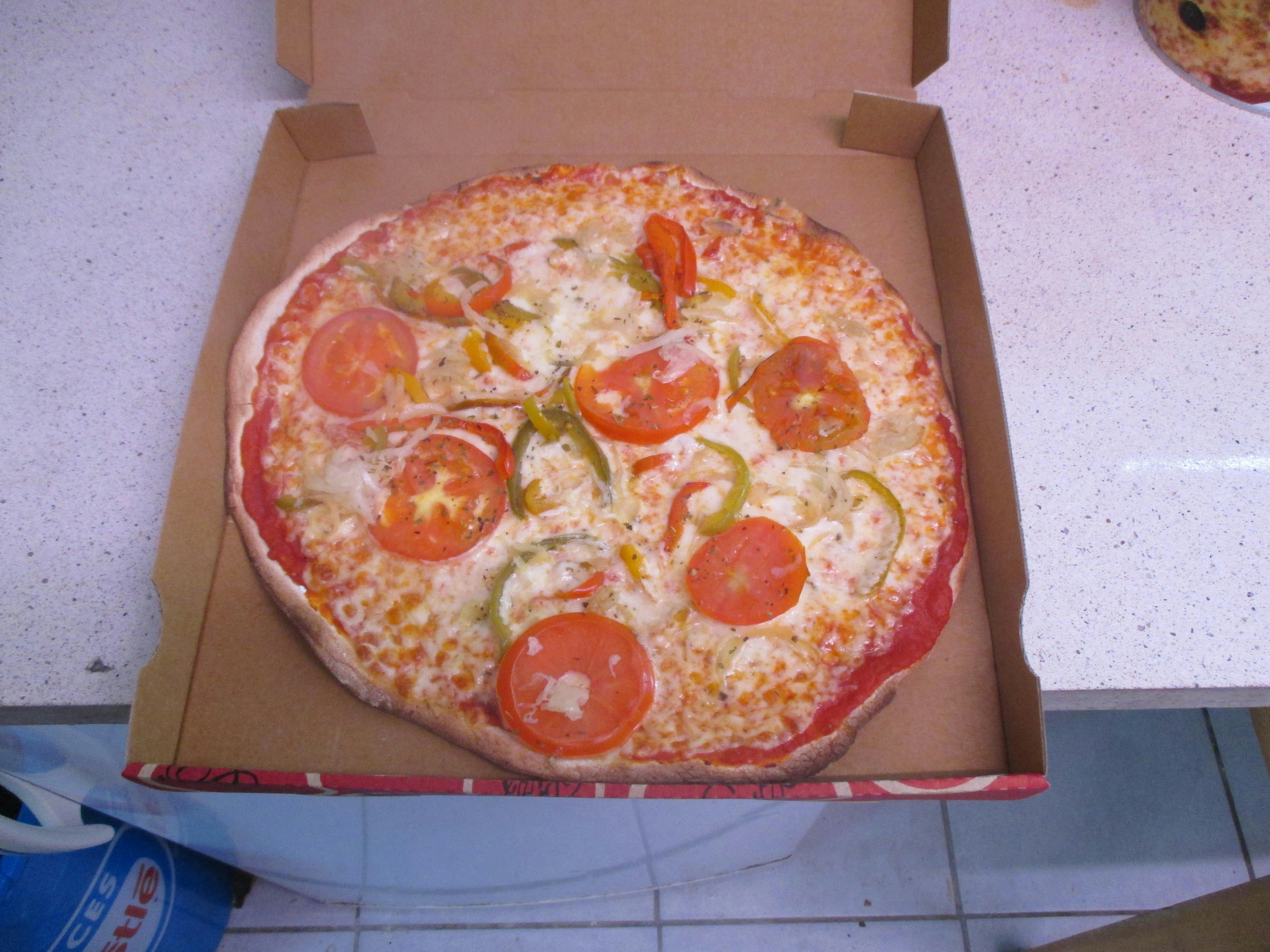 Pizza in einer Kartonschachtel auf einer Fliesenfläche, belegt mit Salami, Paprika und Zwiebeln.
