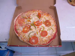 Pizza in einer Kartonschachtel auf einer Fliesenfläche, belegt mit Salami, Paprika und Zwiebeln.