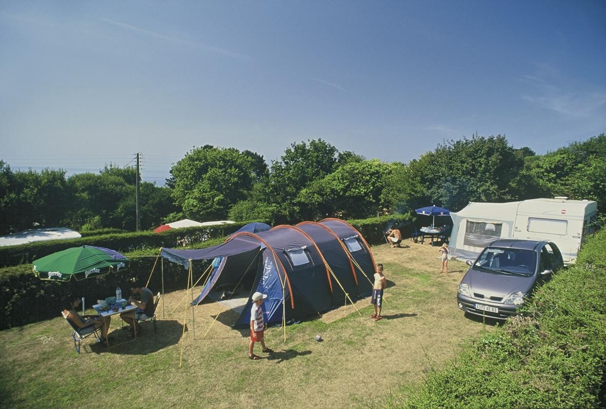 Campingplatz mit mehreren Zelten, einem Wohnwagen und einem Auto auf einer Wiese, daneben ein Tisch und Stühle.