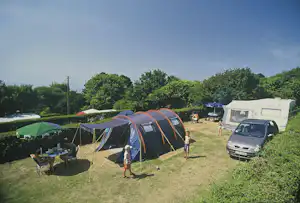 Campingplatz mit mehreren Zelten, einem Wohnwagen und einem Auto auf einer Wiese, daneben ein Tisch und Stühle.