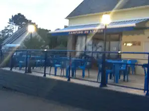Terrasse am Camping de l’Arbre mit blauen Tischen und Stühlen, Gebäude im Hintergrund und Bäume links.