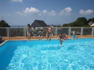 Kinder und Erwachsene springen im Camping-Pool, im Hintergrund Zaun, Liegen und ein Gebäude mit Satteldach.