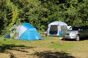 Zwei Campingzelte auf einer Wiese am Stellplatz, daneben ein Auto und ein kleiner Tisch vor dichtem Grün.