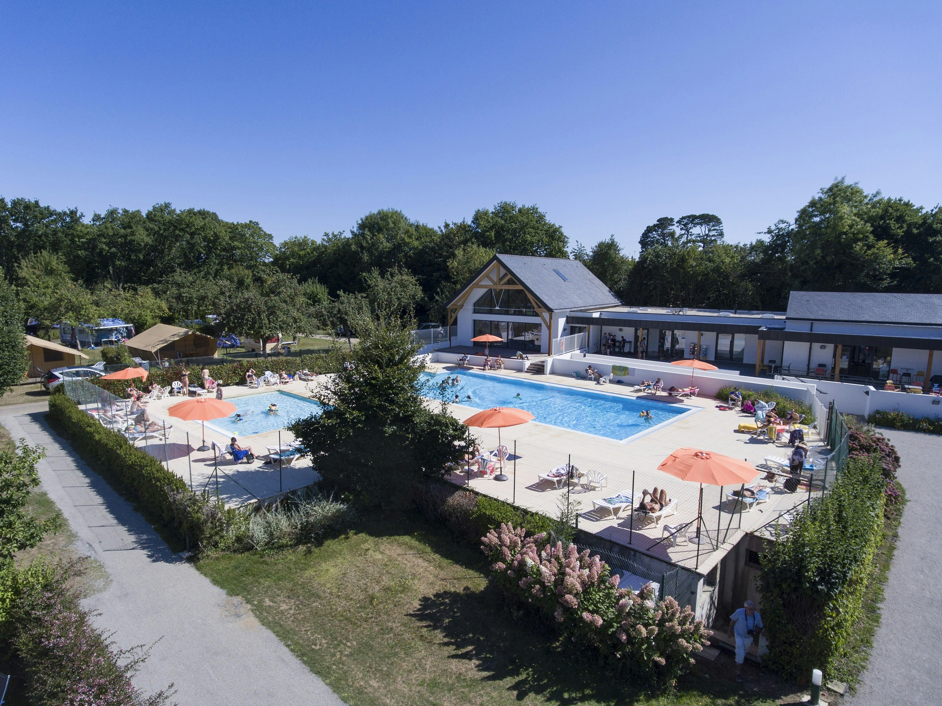 Campingplatz mit Schwimmbecken, Sonnenliegen und Sonnenschirmen, umgeben von Hecken und Bungalows im Hintergrund.