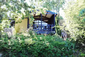 Bungalow mit blauer Veranda, Holzgeländer und Treppe im grünen Garten mit Bäumen und Sträuchern.