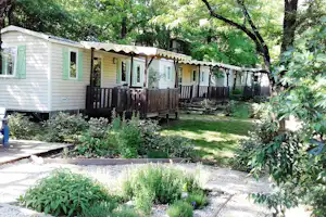 Mobilheim mit Holzterrasse und Markise, umgeben von Rasen, Sträuchern und Bäumen im Campingbereich.