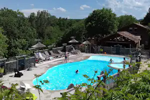 Campingpool mit blauem Wasser, Holzterrasse, Liegestühlen und Sonnenschirmen, umgeben von Bäumen und einem Gebäude.