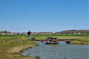Ein Mann steht am Ufer eines kleinen Angelteichs auf dem Campingplatz, mit Grasflächen und Feldern in der Ferne.