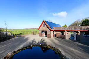 Einfahrt mit gepflastertem Weg und kleinen Teich im Vordergrund, daneben ein Ferienhaus mit Holzverkleidung und blauem Dach.