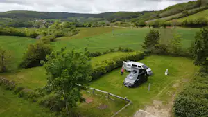 Wohnmobil auf Stellplatz am Rand eines grünen Hügellands, Feldweg und Hecken im Vordergrund, weite Landschaft im Hintergrund.