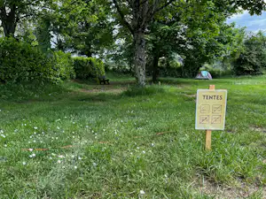 Wiese mit Hinweisschild „TENTES“ und Sitzbank im Grünen, im Hintergrund Zelt in der Nähe von Bäumen.