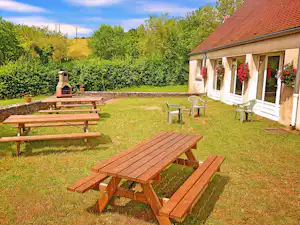 Holz-Tisch und Bänke im Campinggarten vor einer Terrasse mit Blumen und Blick auf Hecken und Bäume.