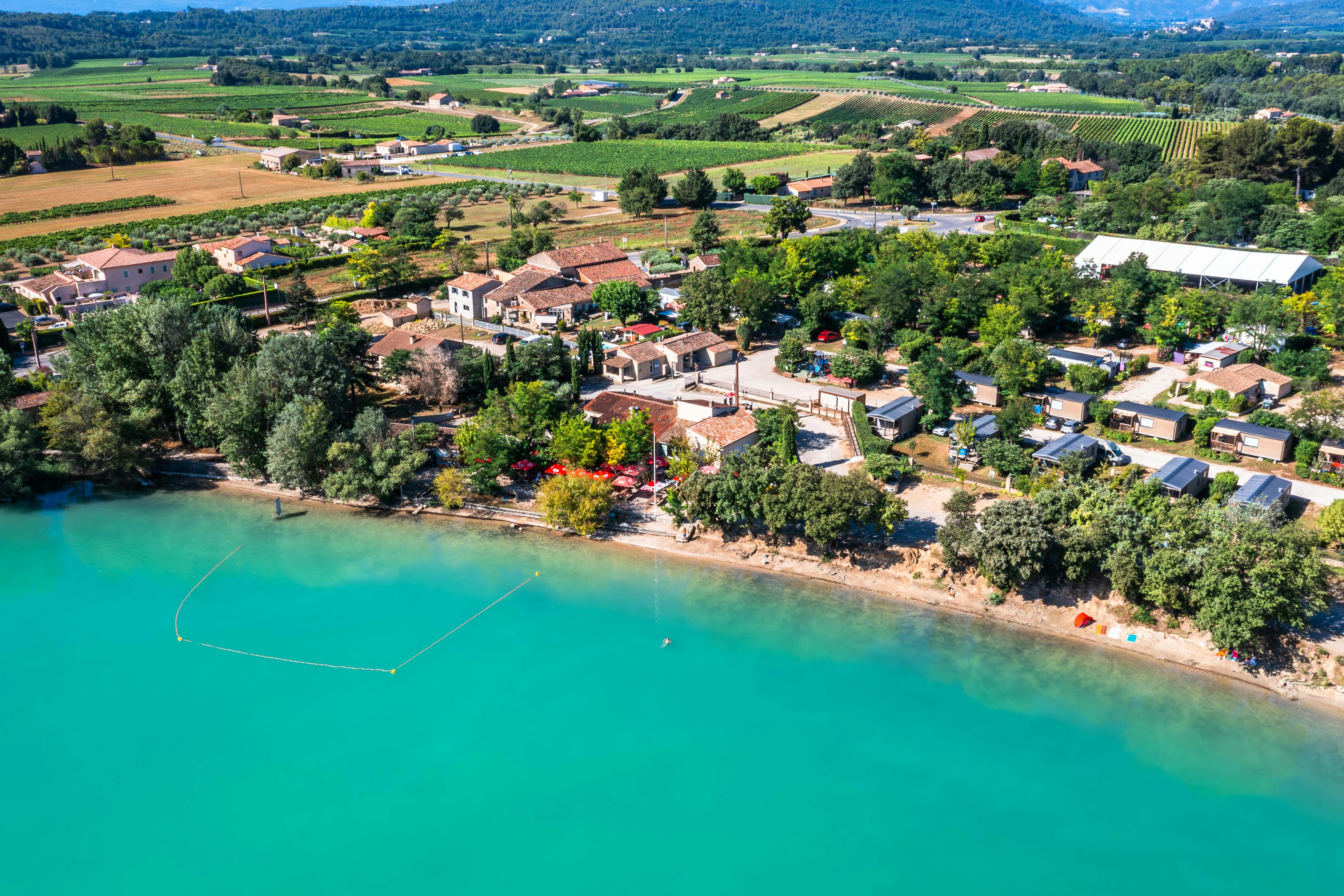 Luftaufnahme vom Campingplatz am türkisfarbenen See mit Ufer, Bäumen, Wegen und vielen Wohnmobilen sowie Zelten.