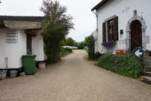 Schmale Gasse zwischen weißen Gebäuden, Hecken und einem Baum, mit Kiesweg und Mülltonnen am Rand.