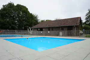 Schwimmbecken mit blauem Wasser und Betonrand vor einem Campinggebäude mit Holzveranda und Zaun.