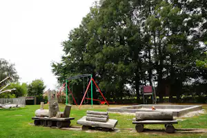 Spielplatz am Campingplatz mit rotem Klettergerüst, Sandkasten und mehreren Holz-Bänken auf dem Rasen.