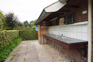 Außenküche am Campingplatz an einer Ziegelwand mit Spüle, Arbeitsplatte und überdachtem Bereich, daneben Hecke und gepflasterter Weg.