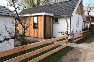 Ferienhaus mit Holzfassade, überdachtem Eingang und Terrasse mit Holzgeländer, im Garten mit Bäumen und Hecken.