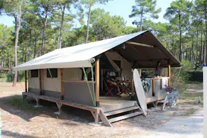 Campingzelt mit Veranda auf Holzpodest, davor Tisch und Stühle, im Hintergrund Kiefern und Waldweg.
