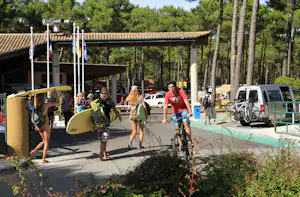Campingplatz-Weg mit Wohnmobilen, Fahrrädern und mehreren Personen vor einem Gebäude, im Hintergrund Bäume und Sonnenschirme.