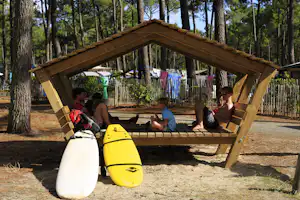 Holzunterstand am Campingplatz im Wald mit Sitzbank, Kajaks und zwei Personen im Hintergrund.