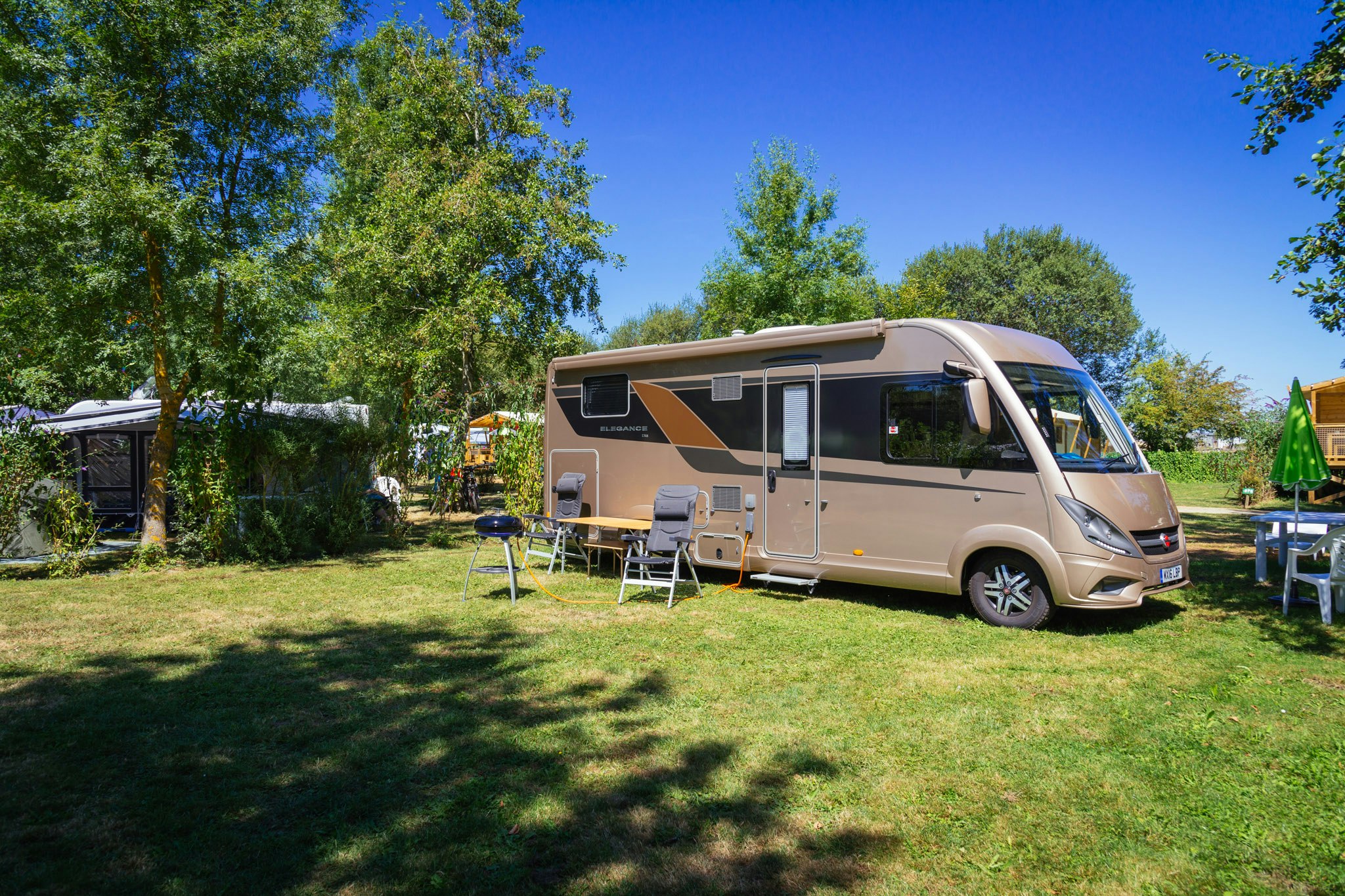 Wohnmobil mit ausklappbarer Markise und Sitzgruppe auf dem Rasen, umgeben von Bäumen und weiteren Stellplätzen.