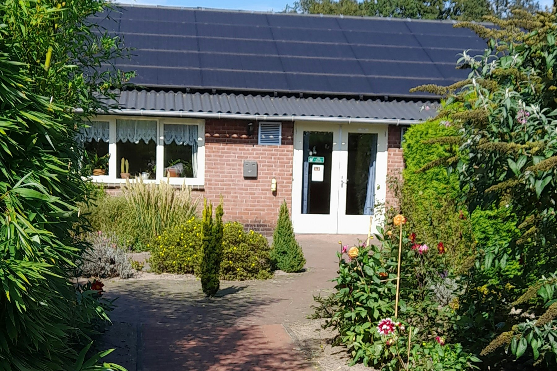 Kleines Ferienhaus mit Ziegelfassade, Eingangstür und Fensterfront, umgeben von Hecken und Blumen im Gartenweg.