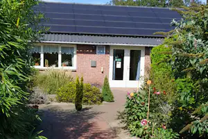 Kleines Ferienhaus mit Ziegelfassade, Eingangstür und Fensterfront, umgeben von Hecken und Blumen im Gartenweg.