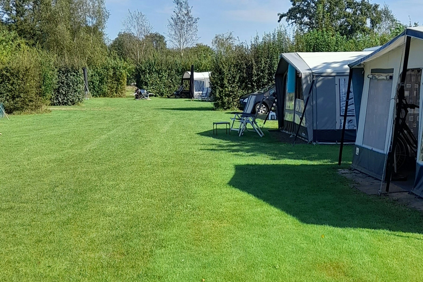 Campingplatz mit großem Rasen, zwei Zelten und Gartenmöbeln; im Hintergrund Bäume und ein weiterer Stellplatz.