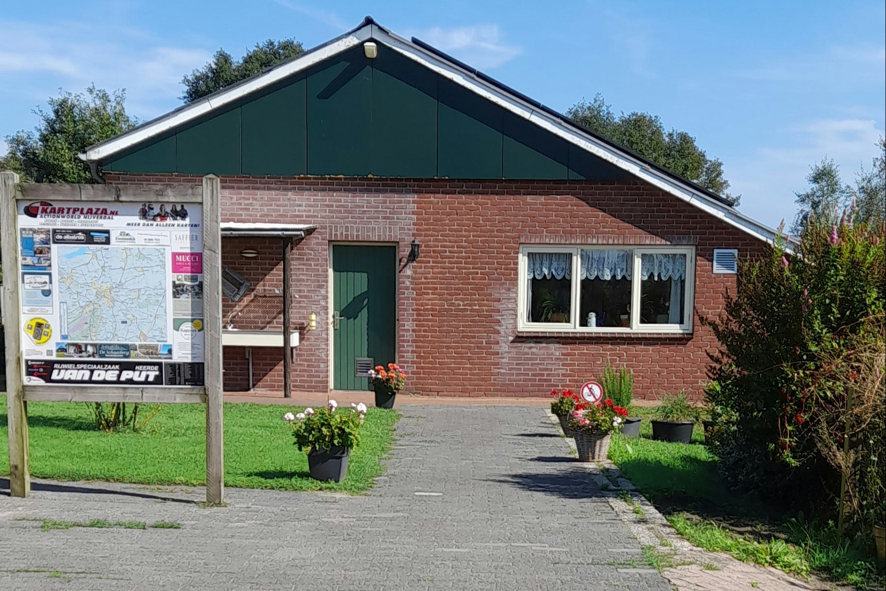 Ziegelbungalow mit grüner Dachfläche, Eingangstür und Fenstern; gepflasterter Weg, Blumen und Töpfe vor dem Gebäude.