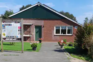 Ziegelbungalow mit grüner Dachfläche, Eingangstür und Fenstern; gepflasterter Weg, Blumen und Töpfe vor dem Gebäude.
