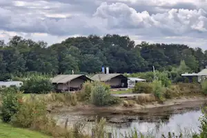 Campingplatz am Fluss mit kleinen Hütten und Zelten, umgeben von Bäumen, Gras und einem Uferweg.