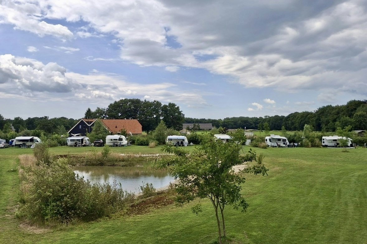 Campingplatz mit Wohnwagen und kleinen Gebäuden, Rasenfläche und Teich im Vordergrund, Bäume und Himmel mit Wolken.