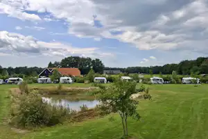 Campingplatz mit Wohnwagen und kleinen Gebäuden, Rasenfläche und Teich im Vordergrund, Bäume und Himmel mit Wolken.