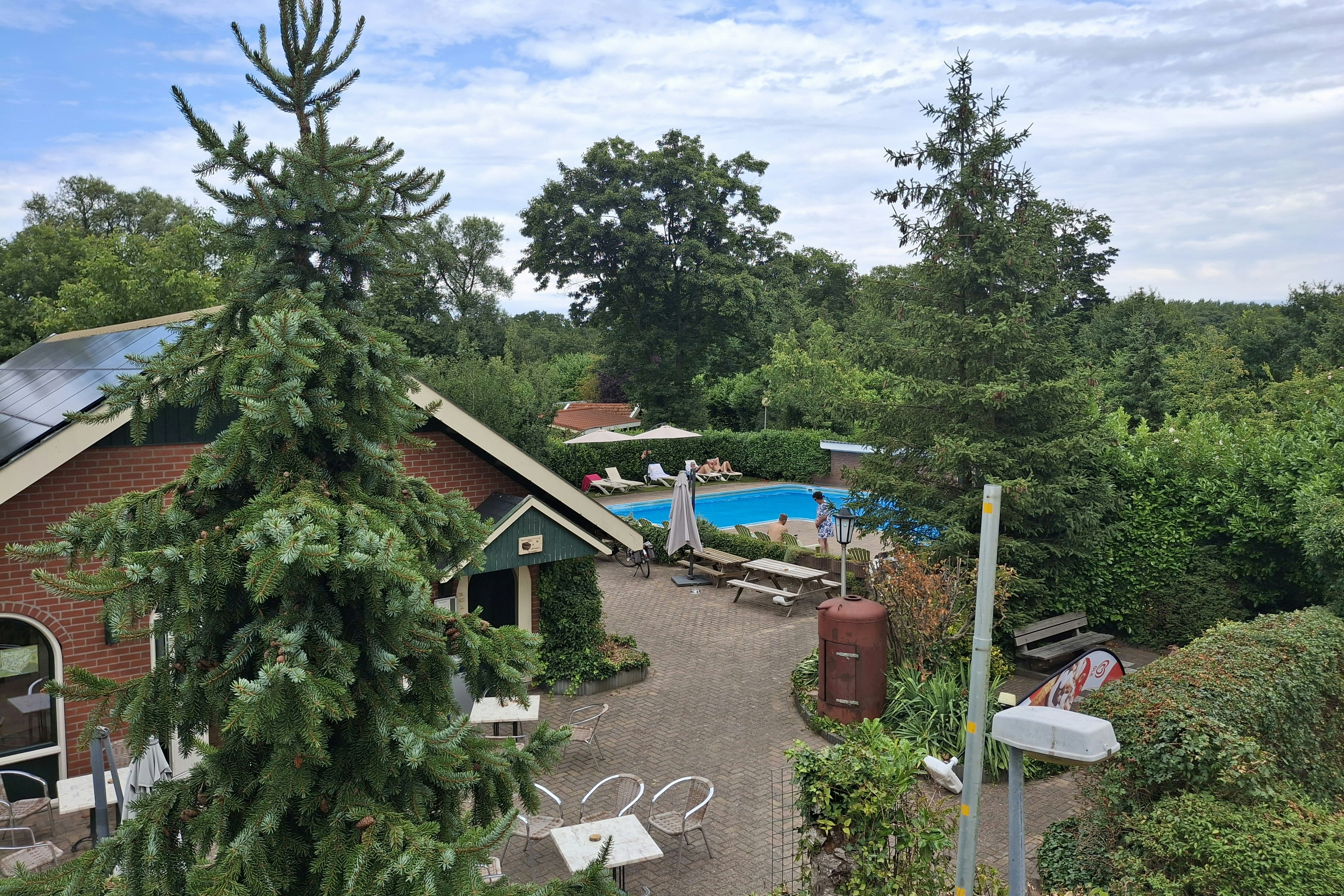 Campingplatz mit kleinen Holzhäusern, Terrasse und Sitzgelegenheiten, im Vordergrund ein Baum, dahinter ein Pool zwischen Bäumen.