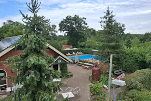 Campingplatz mit kleinen Holzhäusern, Terrasse und Sitzgelegenheiten, im Vordergrund ein Baum, dahinter ein Pool zwischen Bäumen.