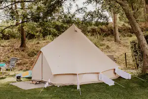 Beiges Zelt auf einer Wiese im Campinggarten, umgeben von Bäumen, mit Campingstuhl und Tisch am Rand.