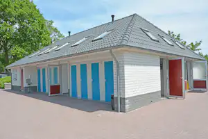 Campinggebäude mit Satteldach, blauen Türen und Ziegelpflaster vor dem Haus, umgeben von Bäumen.